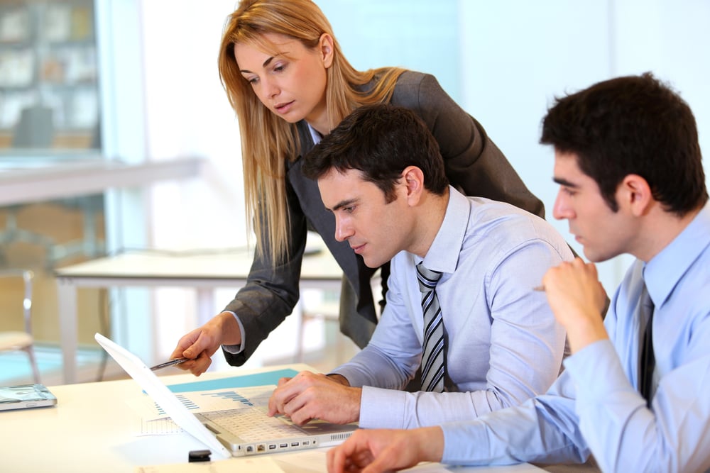 A woman and two men, all in business attire, work together on a project. The woman is pointing at a laptop one man is using while the other man listens intently.