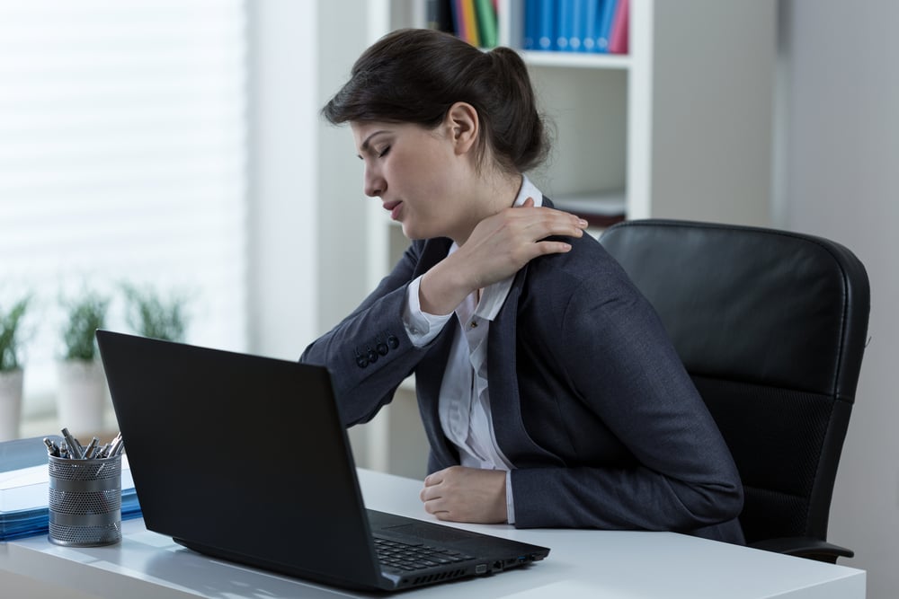 Woman in business attire sitting at a desk grabs her back in pain